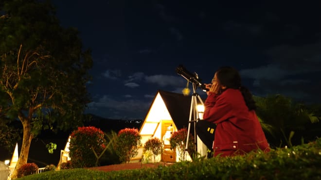 Rheanne looking through a telescope on a lawn at night, lit tents and garden lights behind her.