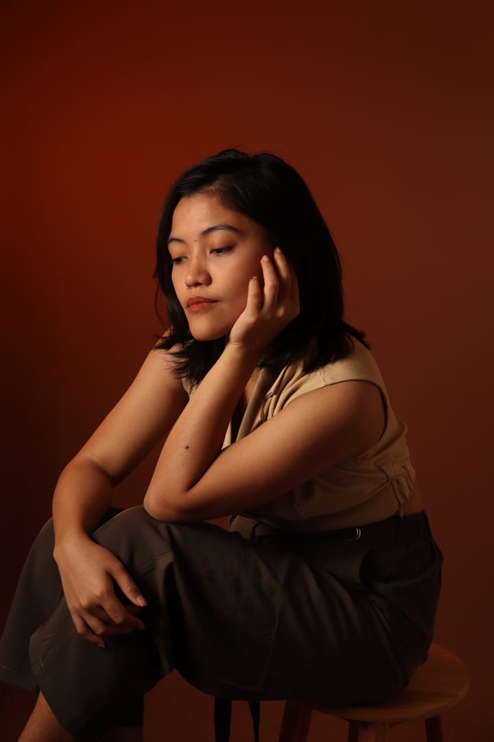 Rheanne seated on a stool against a deep red backdrop, chin resting on one hand.