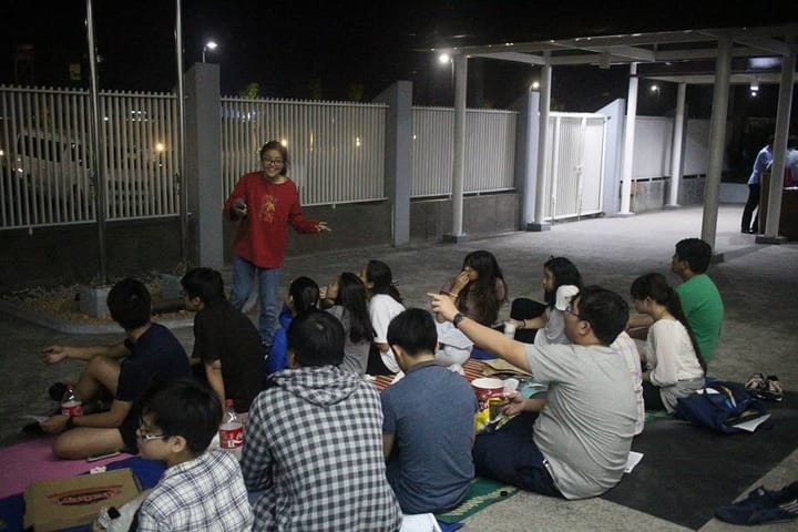 A crowd seated on the ground at an outdoor night stargazing session, a facilitator standing to one side.